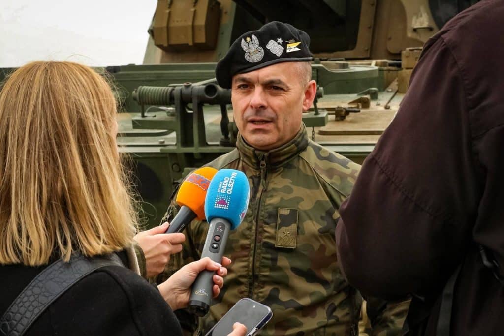 Żołnierz w mundurze i czapce z emblematami, udziela wywiadu dziennikarzom przy nowoczesnym sprzęcie wojskowym na Warmii.