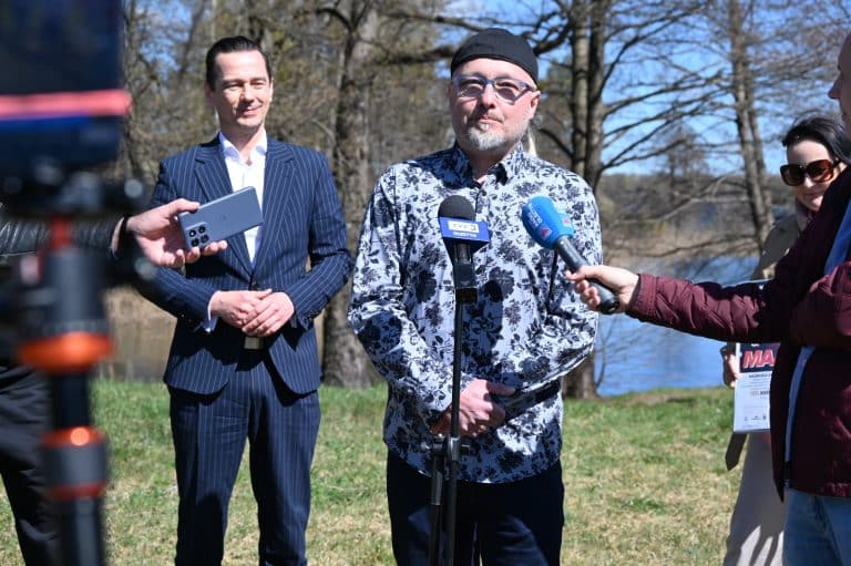Mężczyzna w okularach i czapce, m&oacute;wiący do medi&oacute;w podczas konferencji na świeżym powietrzu, w tle drzewa i jezioro.
