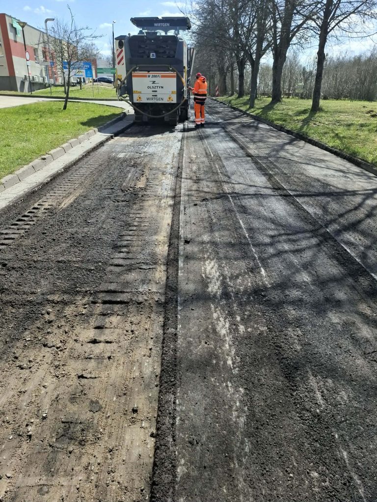 Prace remontowe na ulicy Kurta Obitza w Olsztynie, robotnicy i maszyna na drodze, w tle drzewa i budynki.