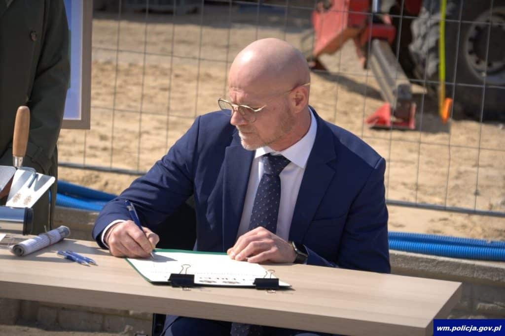 Mężczyzna w garniturze podpisuje dokumenty przy stole na placu budowy nowej komendy policji w Ostr&oacute;dzie.