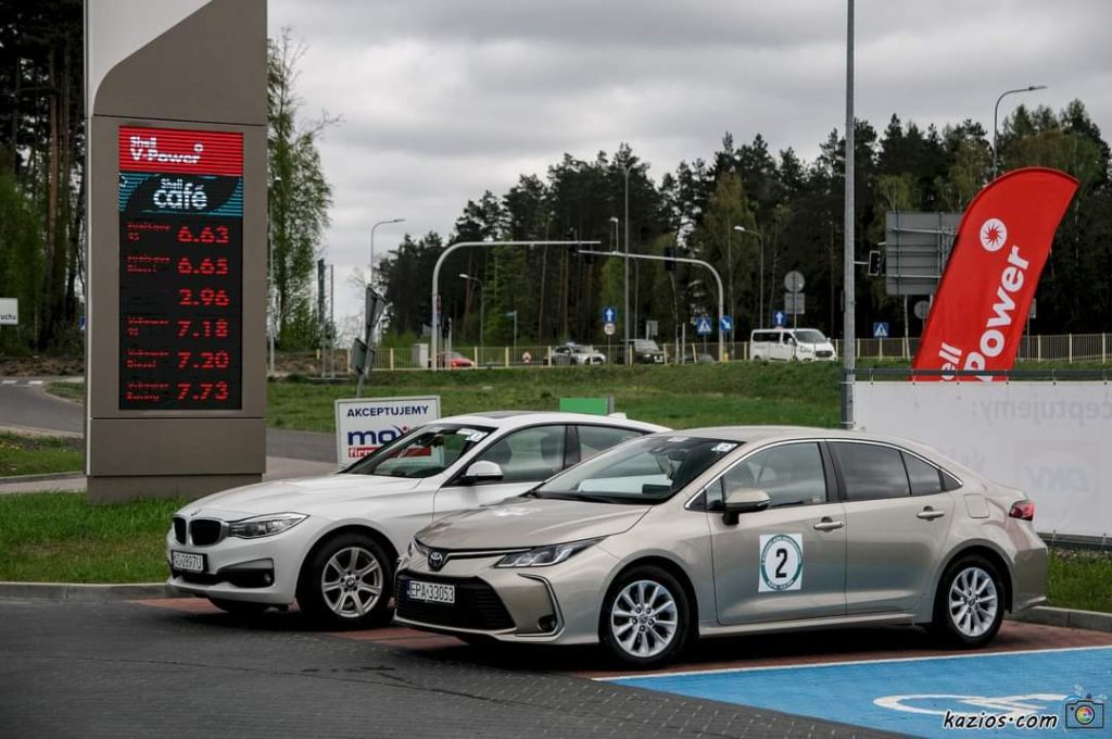 Dwa samochody na parkingu przy stacji paliw z wyświetlaczem cen paliwa w tle.