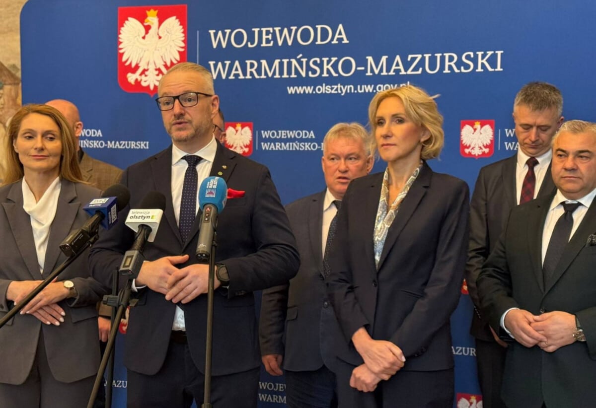 Grupa os&oacute;b na konferencji prasowej przed tablicą z napisem 'Wojewoda Warmińsko-Mazurski'.