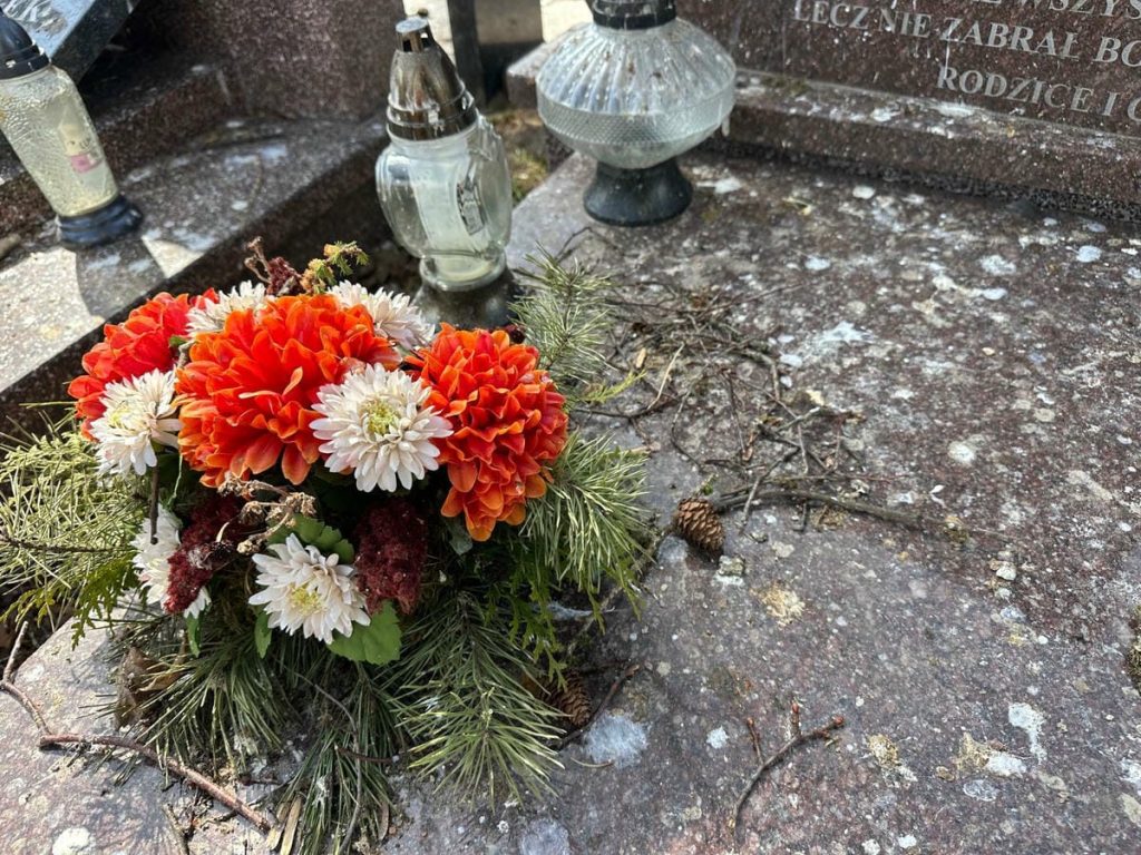 Nagrobek z kwiatami, zanieczyszczony ptasimi odchodami i gałęziami, na cmentarzu w Jezioranach.