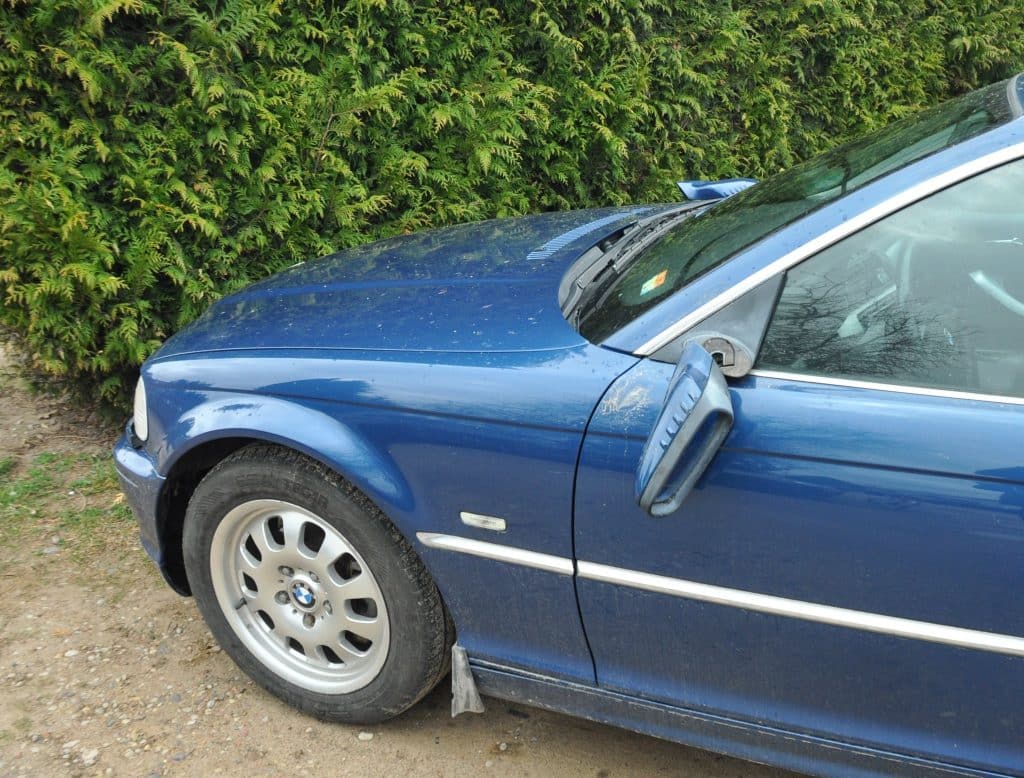 Uszkodzony niebieski samoch&oacute;d BMW z wybitymi lusterkami na tle zielonego żywopłotu.