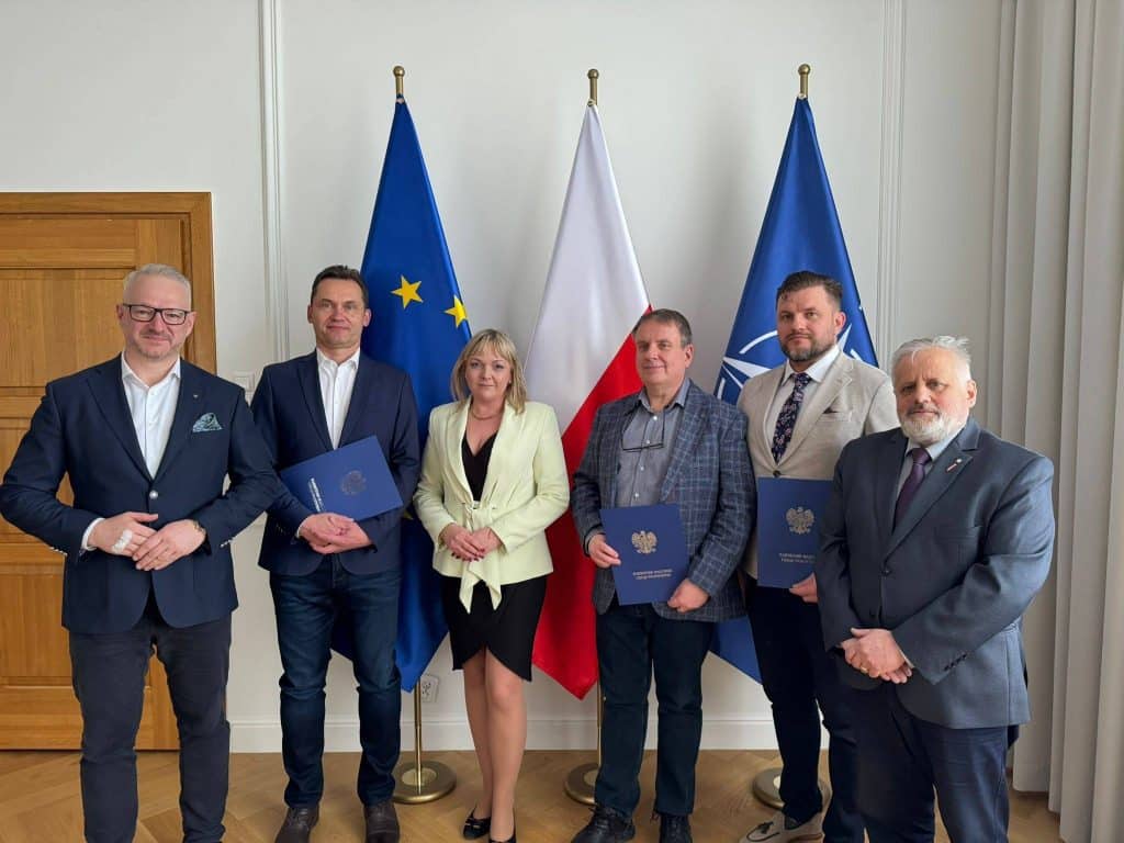 Siedem os&oacute;b na tle flag Unii Europejskiej, Polski i wojew&oacute;dztwa, podczas wręczenia powołań w urzędzie wojew&oacute;dzkim.