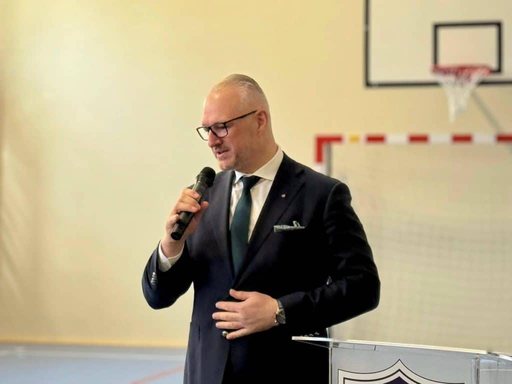 Mężczyzna w garniturze przemawia do mikrofonu na tle boiska sportowego w hali szkolnej.