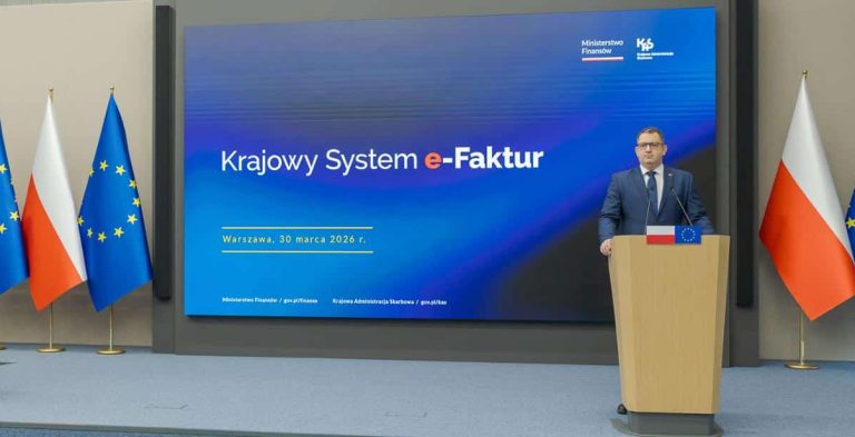 Mężczyzna stoi przy m&oacute;wnicy podczas konferencji o systemie e-Faktur, za nim ekran z informacją o nowym systemie.