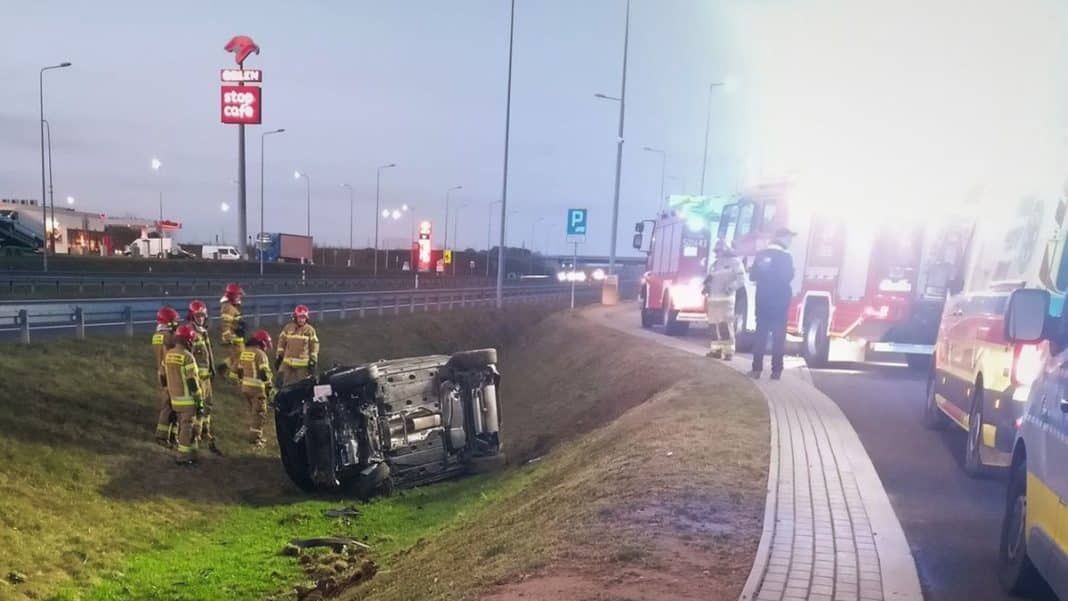 Samoch&oacute;d dachował na poboczu drogi S7, wok&oacute;ł strażacy i pojazdy ratunkowe.