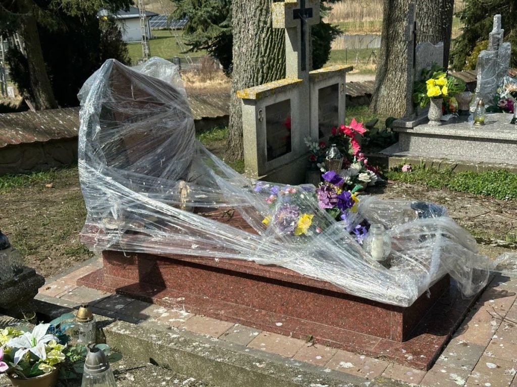 Nagrobek na cmentarzu owinięty folią, wok&oacute;ł kwiaty i znicze, w tle stare nagrobki i drzewa.