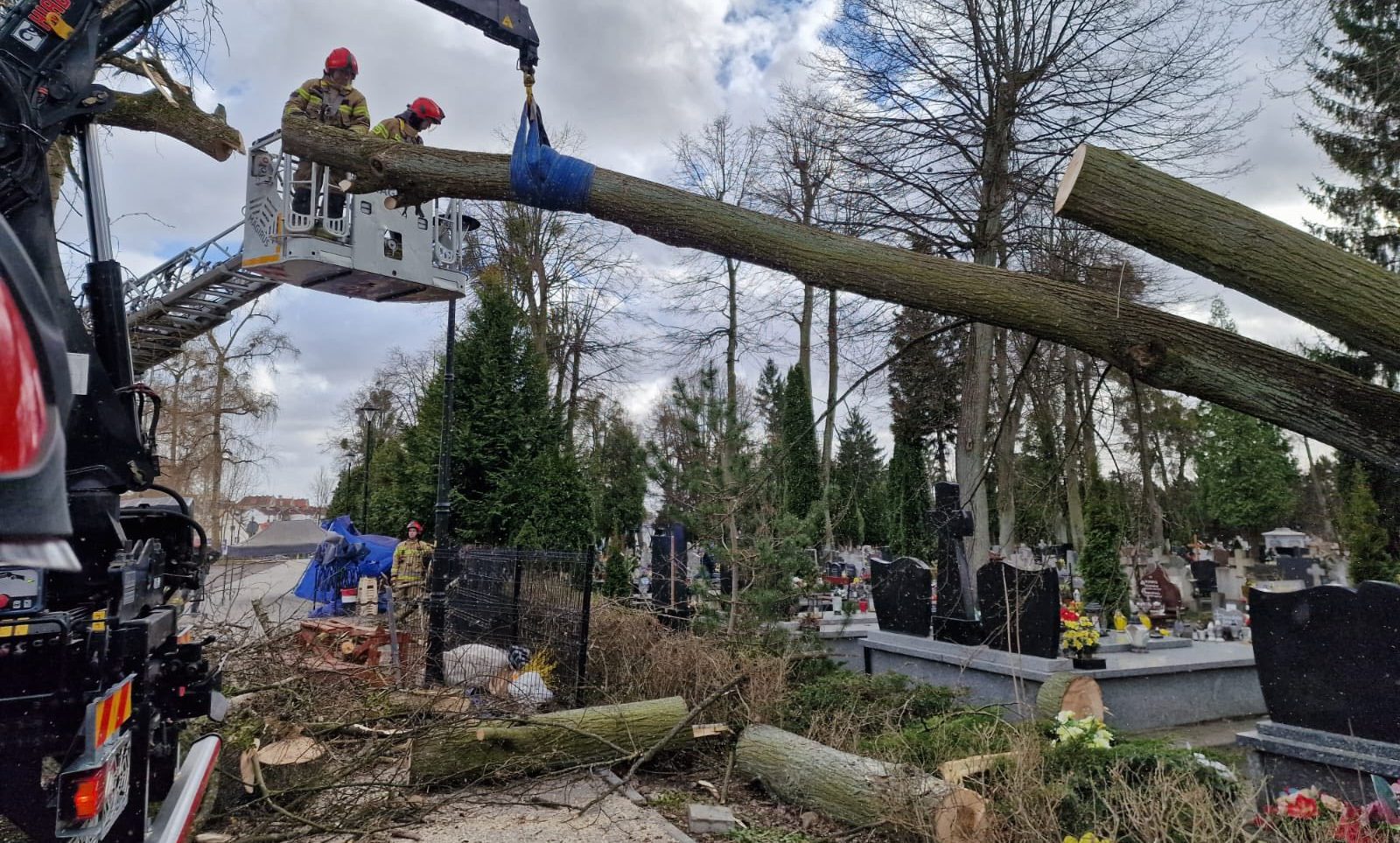 Strażacy usuwają powalone drzewo na cmentarzu po wichurach, w tle widoczne nagrobki i drzewa.