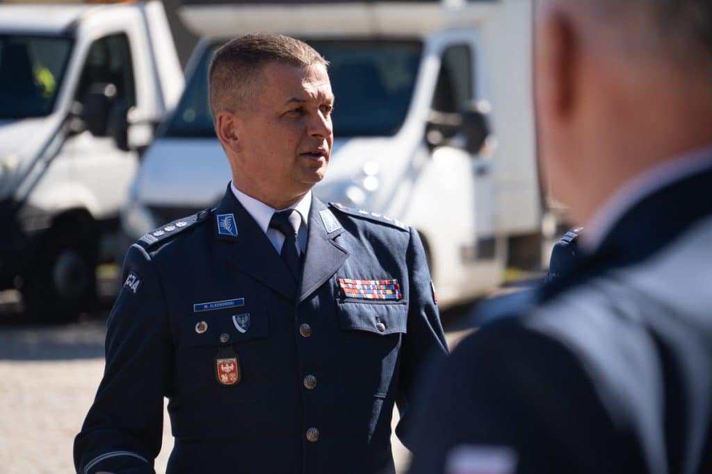 Policjant w uniformie rozmawia z innym funkcjonariuszem na tle policyjnych pojazd&oacute;w na Mazurach.