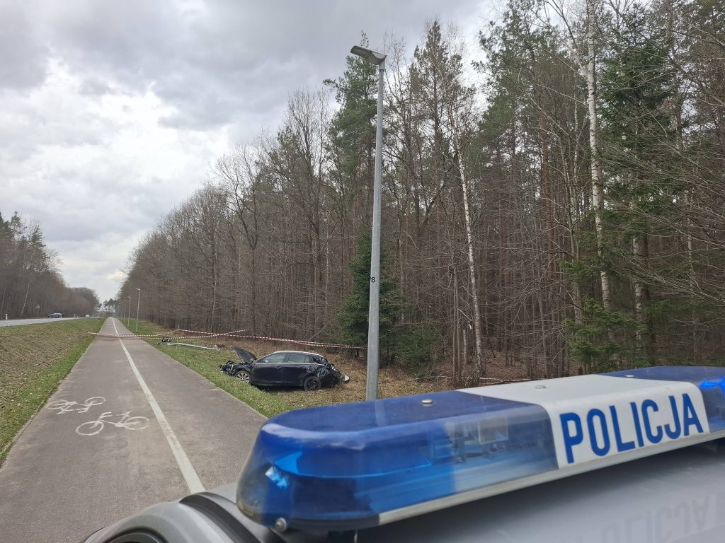 Rozbity samoch&oacute;d na poboczu drogi obok lasu, służby na miejscu zdarzenia, zabezpieczony odcinek trasy.