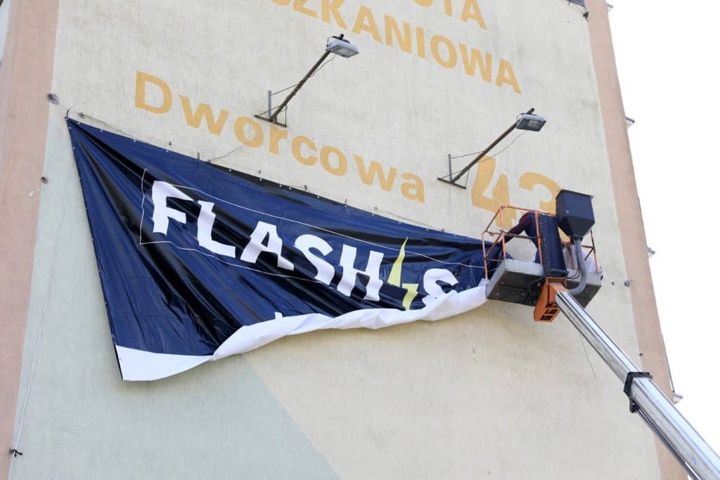 Pracownik w podnośniku montuje baner promocyjny na ścianie budynku w Olsztynie.