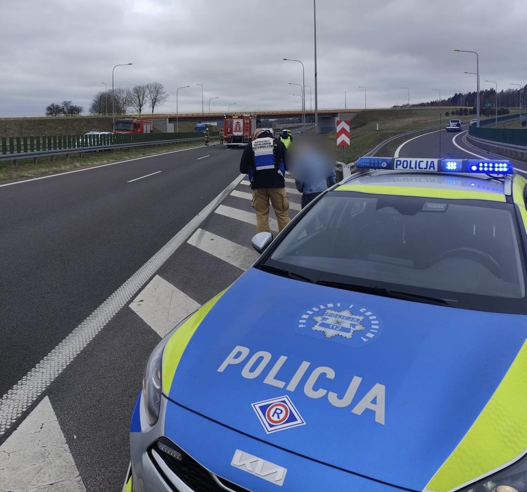 Widok na miejsce dachowania samochodu na trasie S7, z policjantami i służbami ratunkowymi.