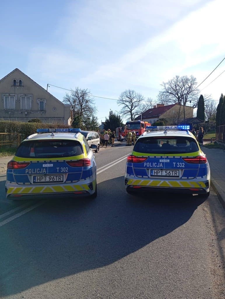 Na zdjęciu widoczne dwa radiowozy policyjne na drodze, w tle akcja ratunkowa i straż pożarna.