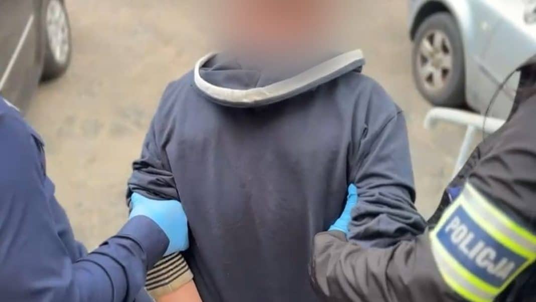 Mężczyzna zatrzymany przez policję, trzymający się za ręce, na tle samochod&oacute;w na parkingu.