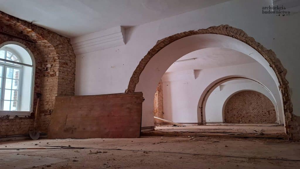 Wnętrze odrestaurowanego koszarowca z cegłami i łukami, trwają prace remontowe.