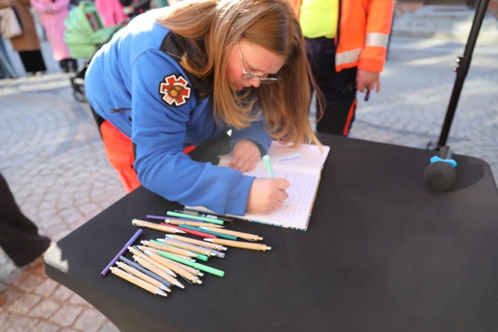 Kobieta zapisuje wspomnienia na tle stołu z wieloma długopisami podczas wydarzenia na Targu Rybnym w Olsztynie.