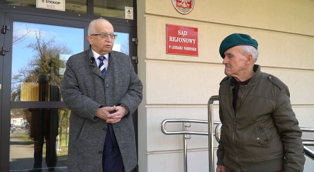 Dw&oacute;ch mężczyzn rozmawia przed sądem rejonowym, jeden w garniturze, drugi w kurtce i beret.