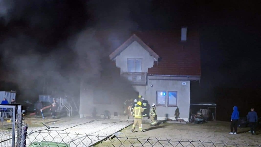 Strażacy gaszą pożar domu jednorodzinnego w nocy, w tle widoczny dym i płomienie.