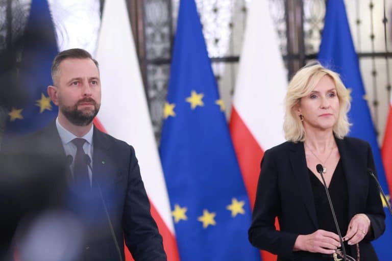 Dwoje polityk&oacute;w stoi przy mikrofonach, za nimi flagi Polski i Unii Europejskiej.