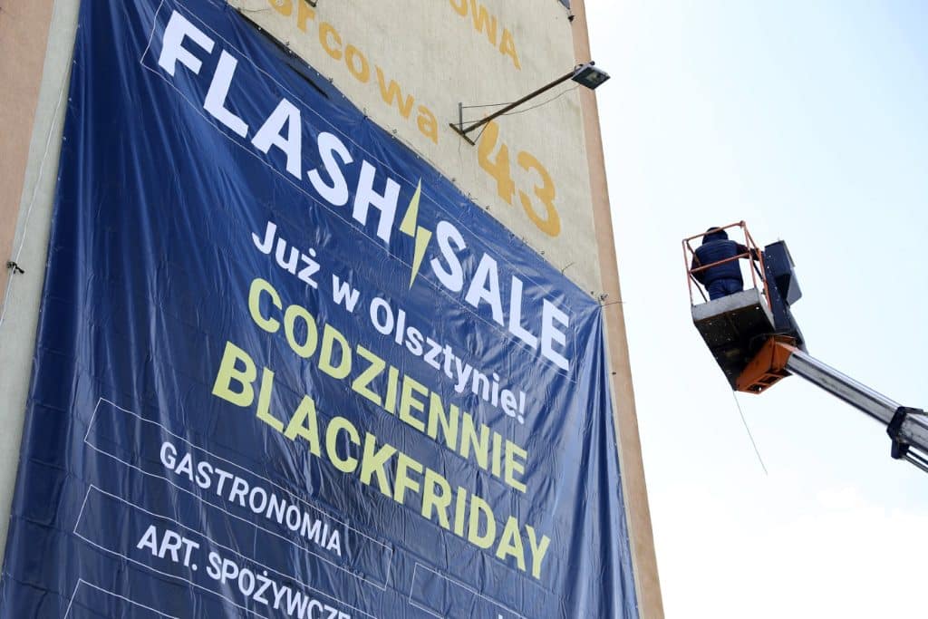 Duży baner promujący platformę Flashsale.pl na budynku w Olsztynie, z pracownikiem na podnośniku montującym reklamę.