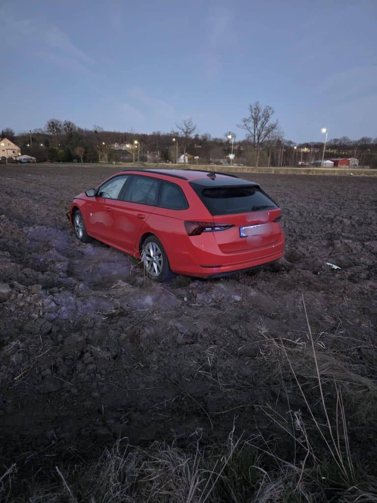 Czerwony samoch&oacute;d na polu po wypadku, zniszczony z tyłu, w okolicy pola i drzew.