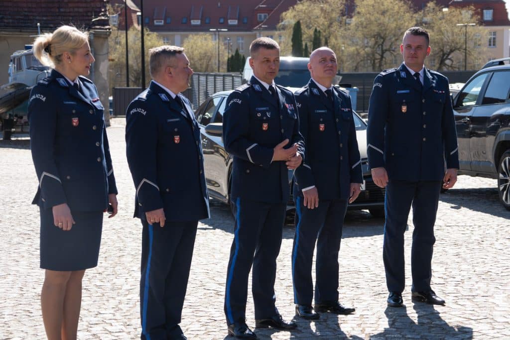Grupa policjant&oacute;w w uniformach stojąca na placu z nowymi radiowozami w tle.