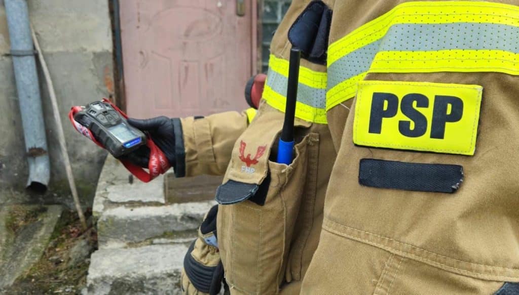 Strażak w mundurze PSP trzyma urządzenie pomiarowe podczas akcji ratunkowej przy wejściu do mieszkania.