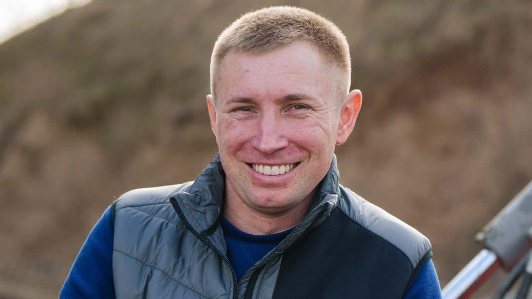 Uśmiechnięty mężczyzna na tle natury, w casualowym ubraniu, na zewnątrz.