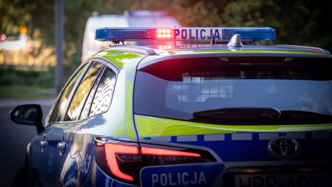 Policja na ulicy, samoch&oacute;d policyjny z włączonymi światłami, w tle drzewa i budynki.