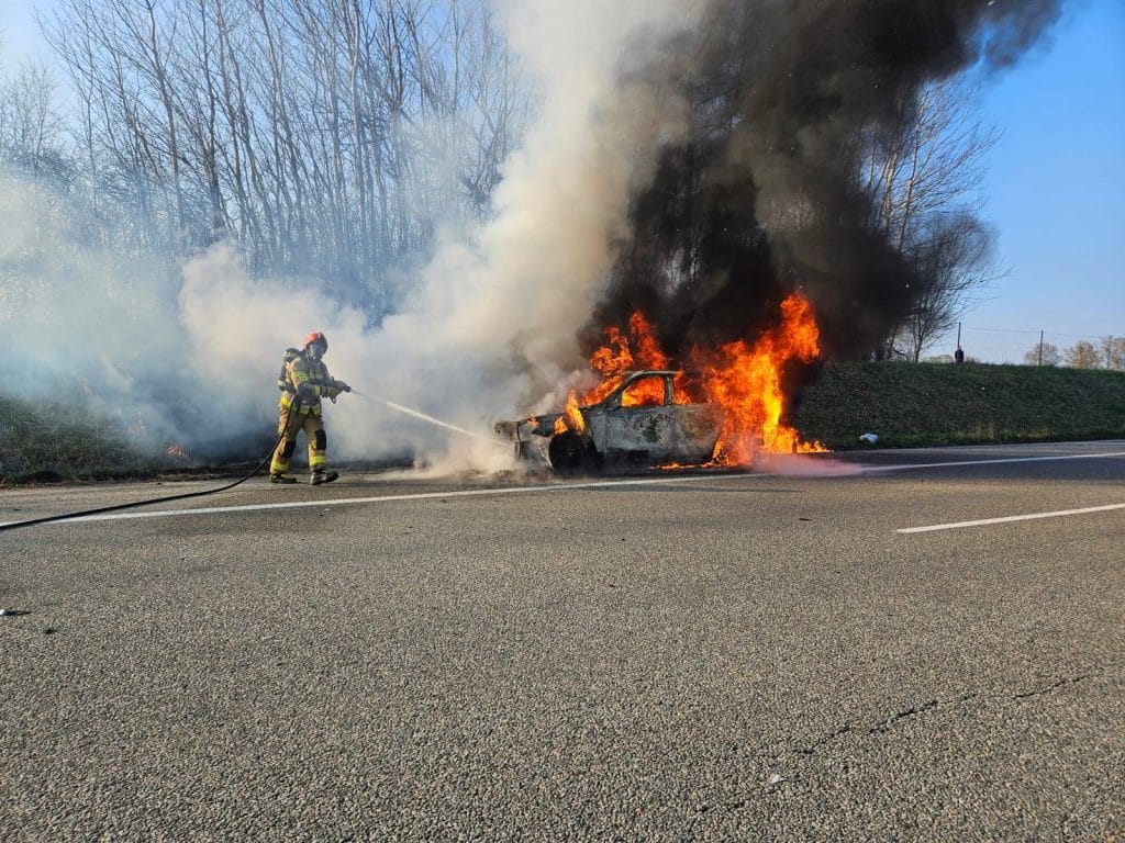 Strażacy gaszą pożar samochodu marki Audi na drodze ekspresowej S22, wok&oacute;ł unoszą się kłęby dymu i ogień.