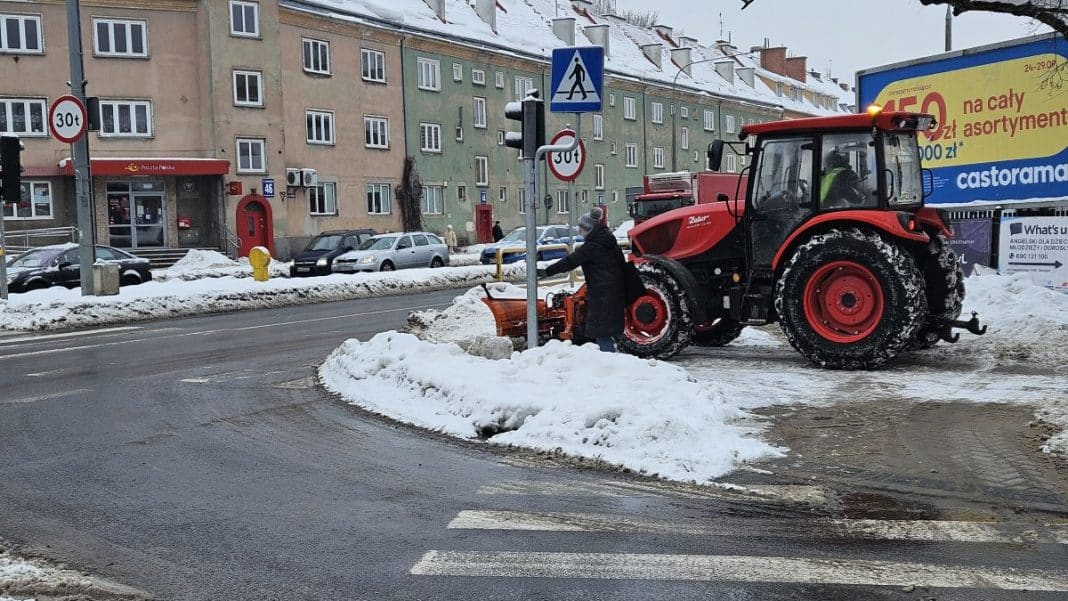 Pracownik odśnieża ulicę w Olsztynie, obok traktor z pługiem, w tle budynki i samochody, zimowa sceneria.