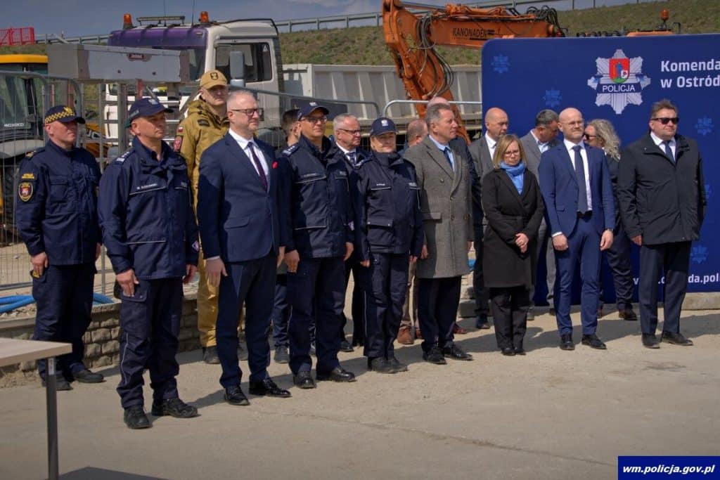 Grupa policjant&oacute;w i urzędnik&oacute;w na placu budowy nowej komendy w Ostr&oacute;dzie, w tle maszyny budowlane.