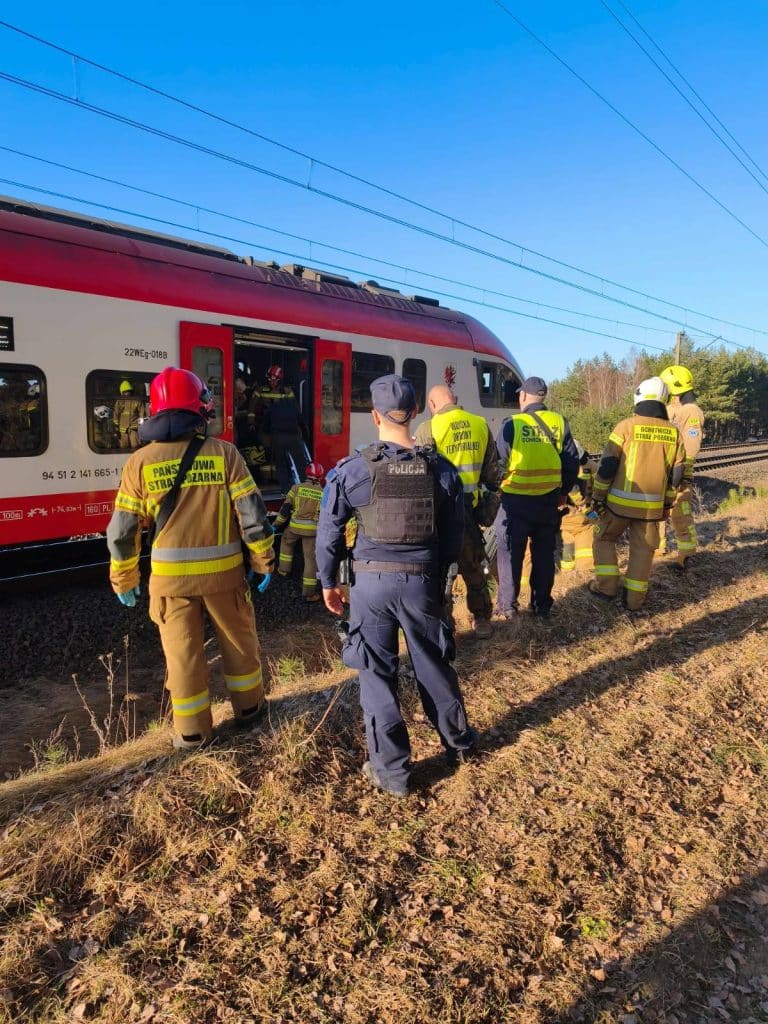 Strażacy i policja na miejscu wypadku na niestrzeżonym przejeździe kolejowym w Lipinkach, obok pociągu i samochodu.