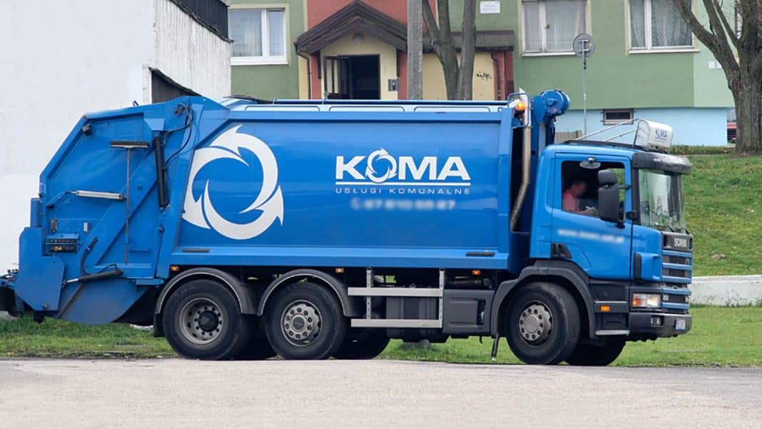 Niebieska śmieciarka podczas odbioru odpad&oacute;w na tle budynk&oacute;w i drzew.