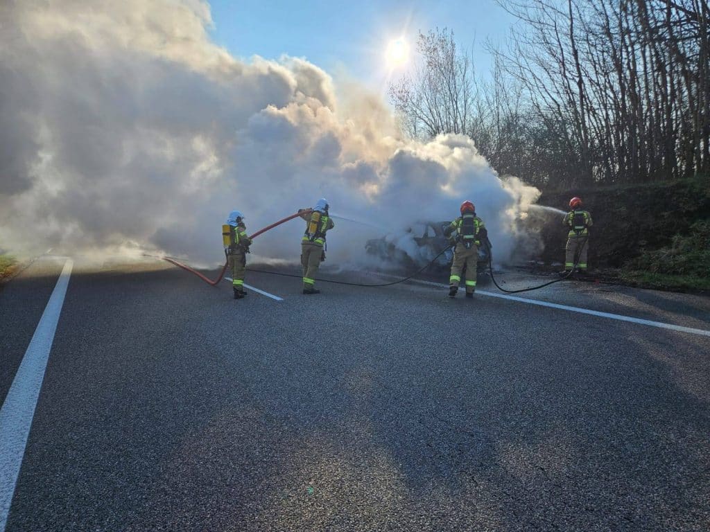 Strażacy gaszą pożar samochodu na drodze ekspresowej S22, w tle drzewa i słońce.