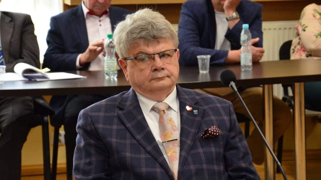 Mężczyzna w garniturze z krawatem siedzi przy stole podczas spotkania, w tle inni uczestnicy.