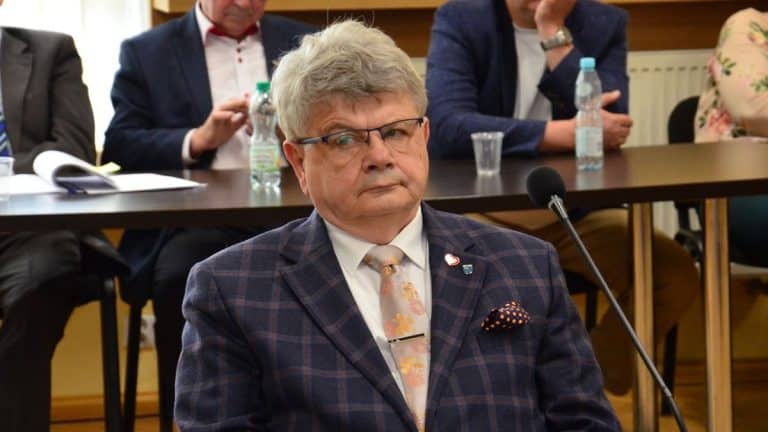 Mężczyzna w garniturze z krawatem siedzi przy stole podczas spotkania, w tle inni uczestnicy.