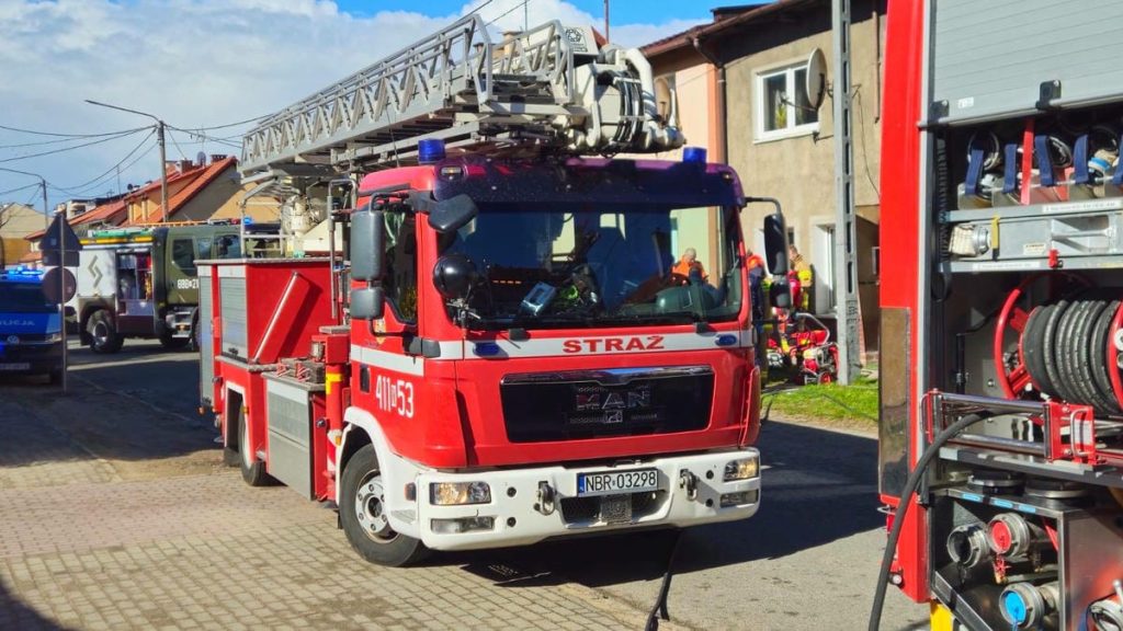 Straż pożarna na miejscu zdarzenia, w tle budynki mieszkalne, w akcji ratunkowej.