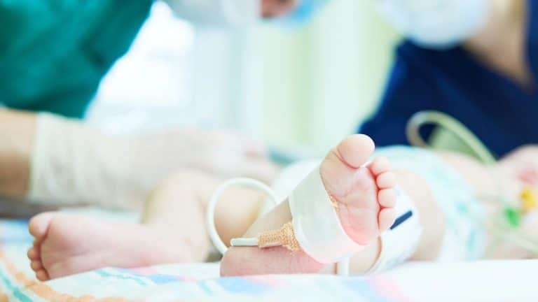 Dziecko w szpitalu z założonymi rurkami i opatrunkami na nogach, leżące na ł&oacute;żku.