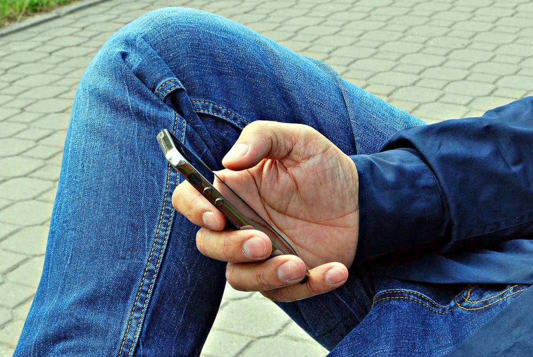Osoba korzysta z telefonu, siedząc na zewnątrz na brukowanej powierzchni, w jeansach i kurtce.