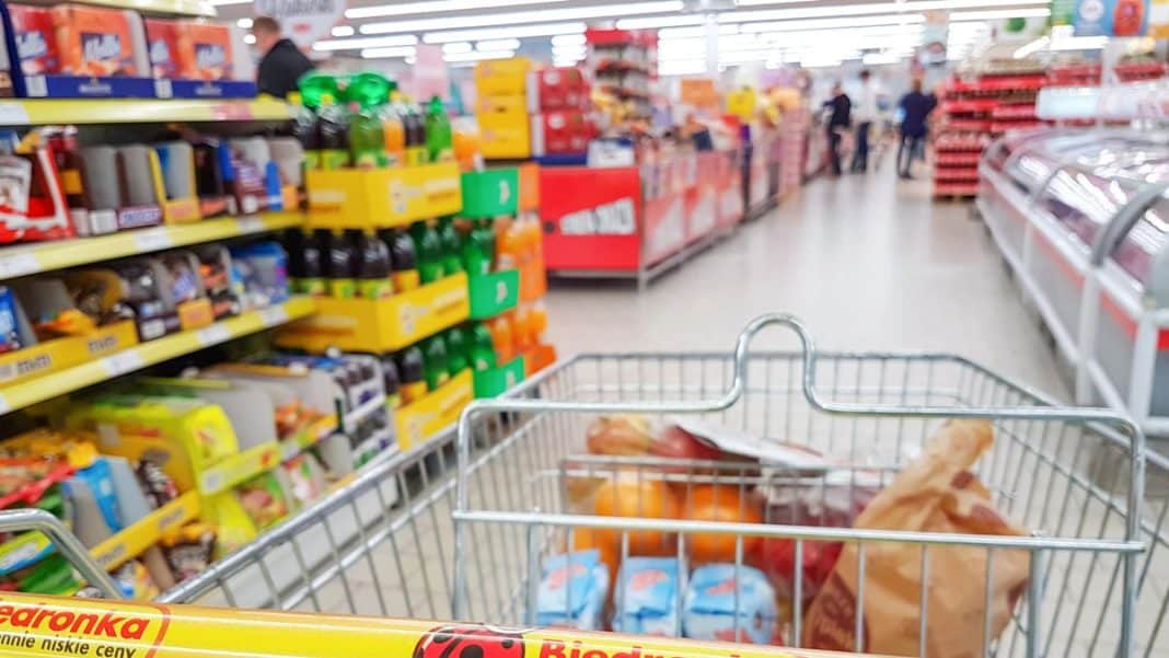 Wnętrze supermarketu z koszykiem pełnym zakup&oacute;w na tle p&oacute;łek z produktami.