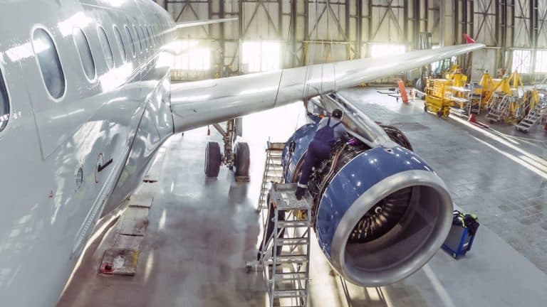 Samolot w hangarze, widoczny silnik i część kadłuba, pracownicy przy obsłudze samolotu.