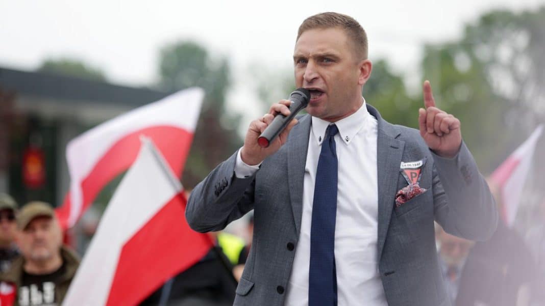 Mężczyzna w garniturze przemawia do tłumu z mikrofonem, w tle flagi i ludzie na zewnątrz.