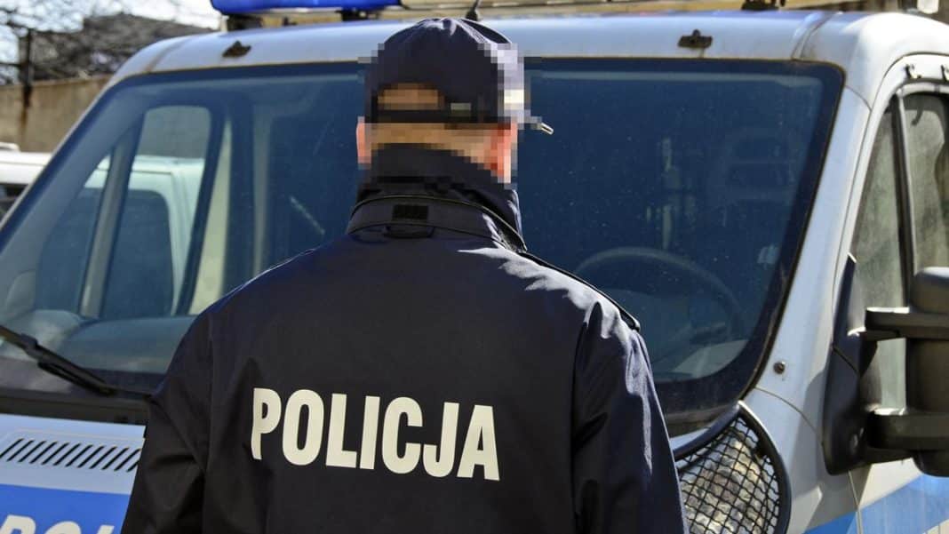 Policjant z napisem 'Policja' na plecach stoi przed radiowozem na Mazurach.