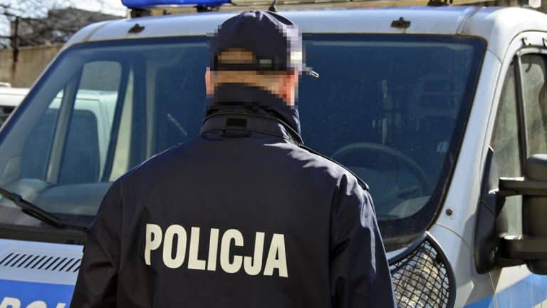 Policjant z napisem 'Policja' na plecach stoi przed radiowozem na Mazurach.