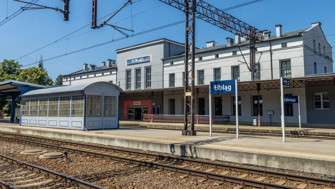 Widok na peron kolejowy w Elblągu z oznaczeniem stacji i torami, słoneczny dzień.