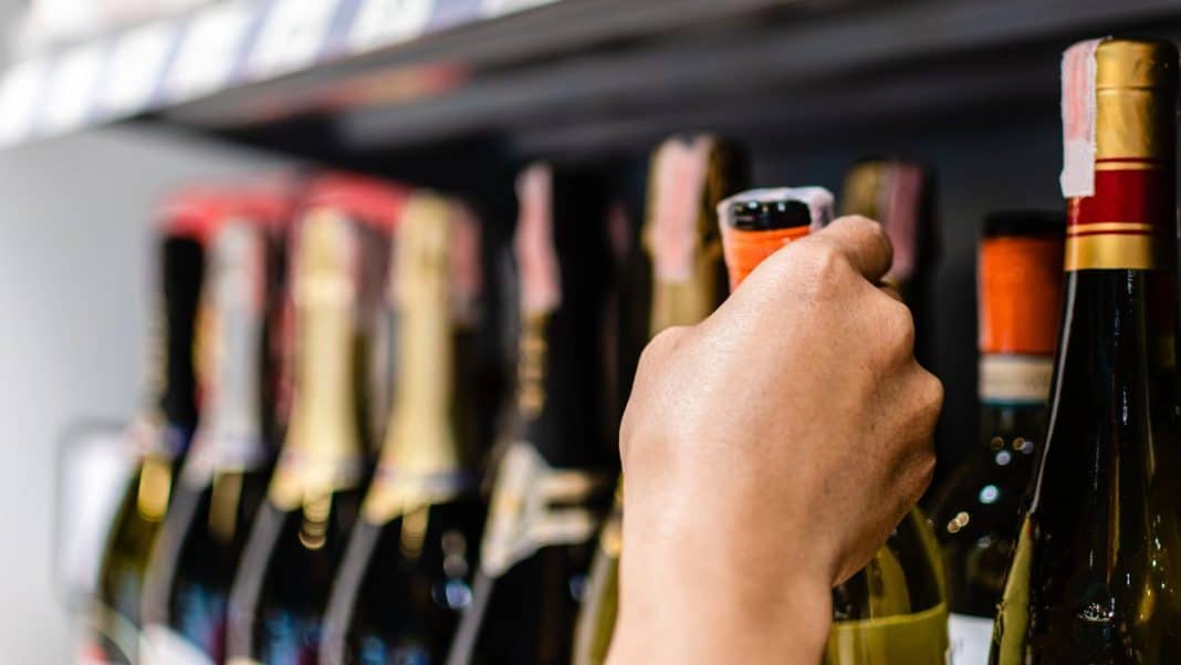 Osoba sięga po butelkę alkoholu na p&oacute;łce w sklepie, widoczne r&oacute;żne butelki napoj&oacute;w alkoholowych.