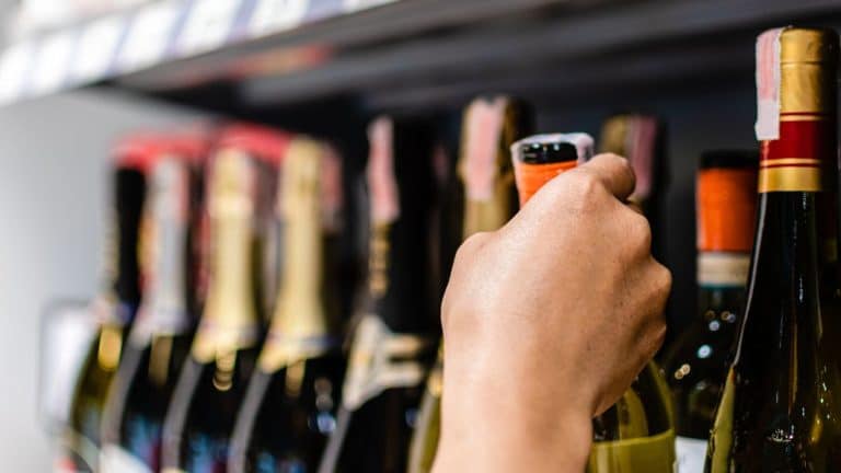 Osoba sięga po butelkę alkoholu na p&oacute;łce w sklepie, widoczne r&oacute;żne butelki napoj&oacute;w alkoholowych.